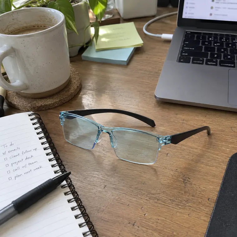 Blue clear frame glasses on a work desk beside notebook, laptop, and coffee
